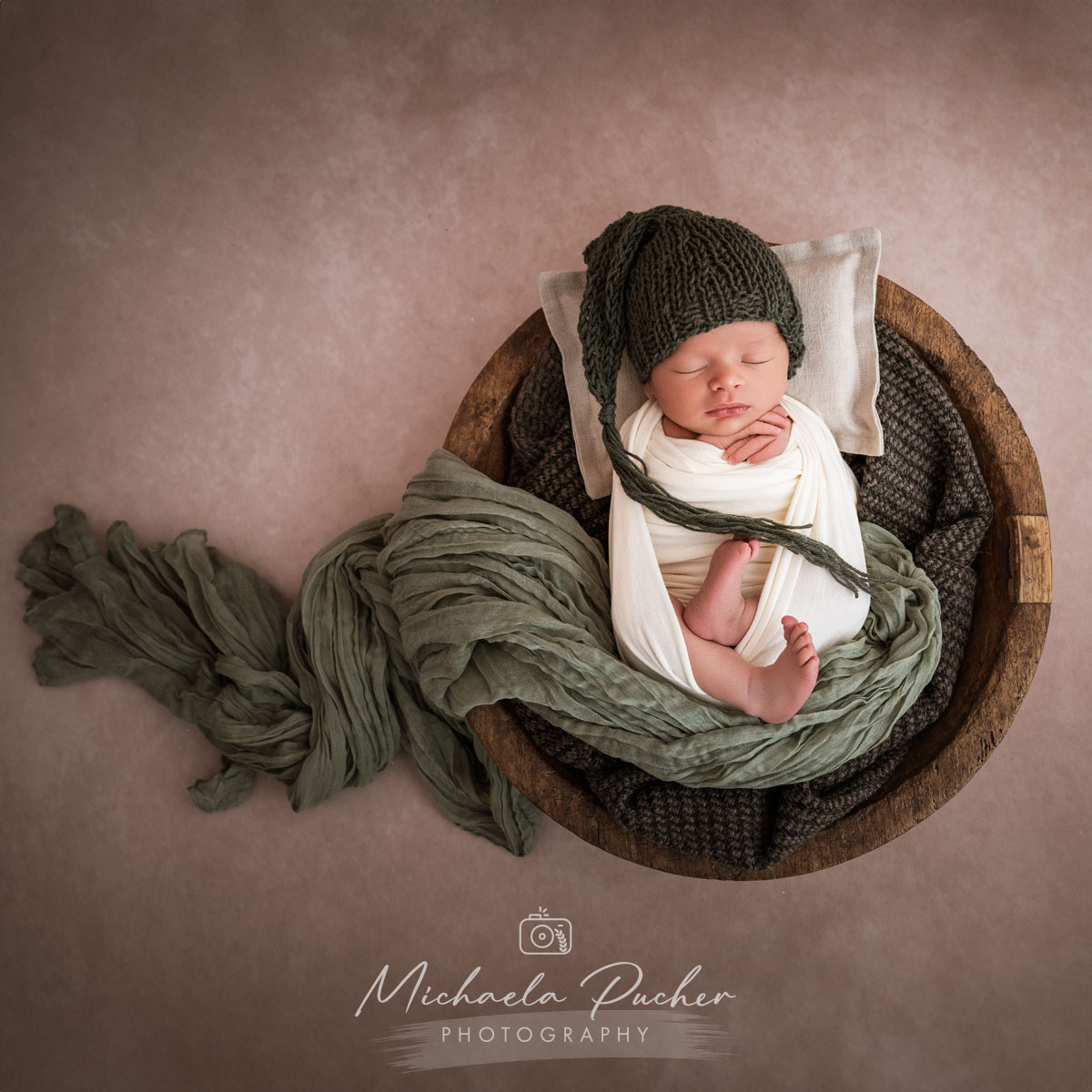 Schlafendes Neugeborenes, eingewickelt in helle Tücher, liegt in einer runden Holzschale; das Baby trägt eine dunkelgrüne Strickmütze und ist von weichen Stoffen in Naturtönen umgeben.